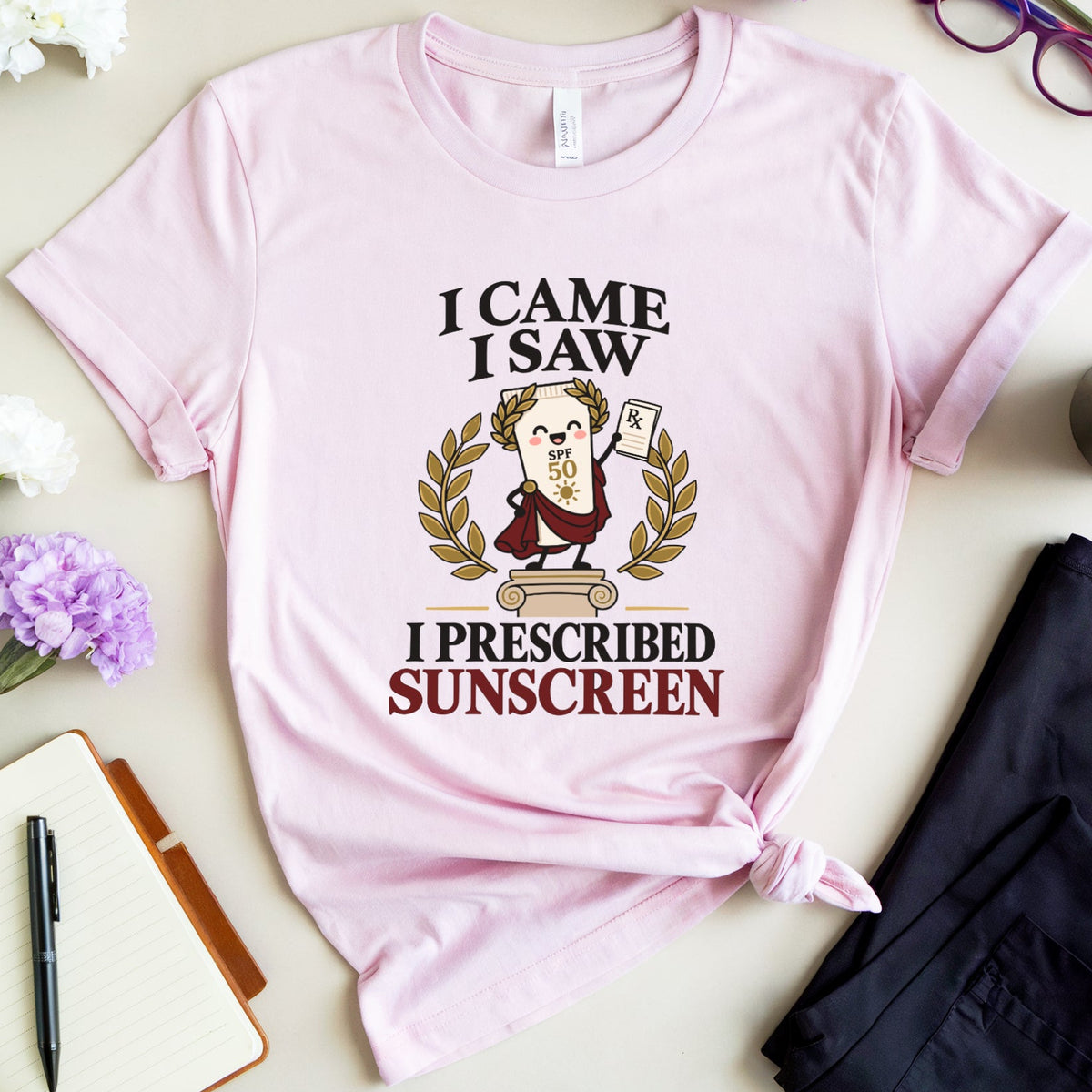 A light pink graphic t-shirt is displayed in a styled flat-lay arrangement on a beige surface. The shirt features a cartoon illustration of a doctor holding a thermometer and a dollar bill, surrounded by text that reads 'I CAME I SAW I PRESCRIBED SUNSCREEN'.