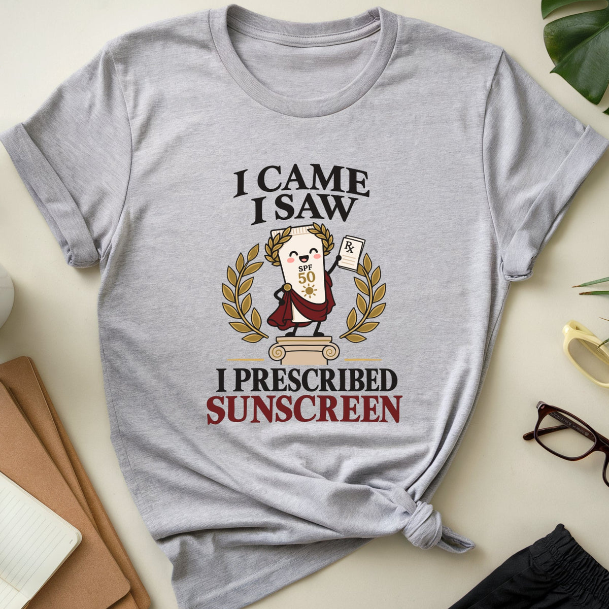A light heather gray graphic t-shirt is displayed in a flat-lay arrangement on a light beige surface. The shirt features a cartoon illustration of a sun character with the text 'I CAME I SAW I PRESCRIBED SUNSCREEN'.