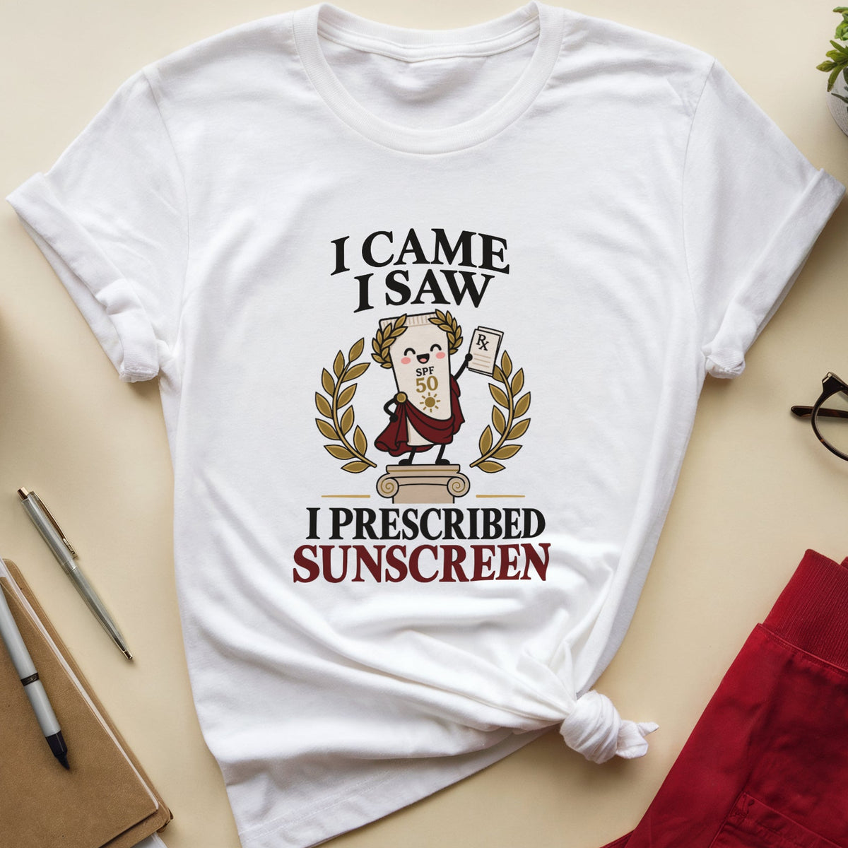A white graphic t-shirt featuring a cartoon illustration of a doctor prescribing sunscreen, with the text 'I CAME I SAW I PRESCRIBED SUNSCREEN'. The shirt is styled in a flat-lay arrangement on a beige background alongside a notebook, pen, coffee cup, glasses, and red pants.