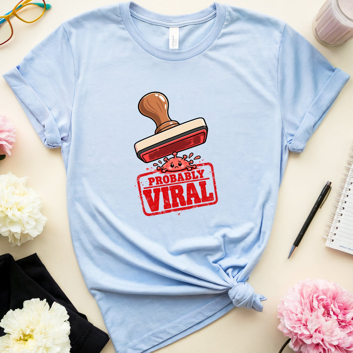 A light blue graphic t-shirt with a 'PROBABLY VIRAL' stamp design, styled in a flat lay with pink and white flowers, glasses, a notebook, and a drink on a beige background.