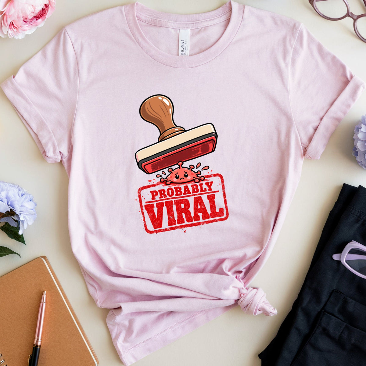 A light pink short-sleeve graphic T-shirt laid flat on a cream surface, surrounded by decorative props including flowers, glasses, a notebook, pens, and folded black pants. The shirt has a large stamp graphic with the words “PROBABLY VIRAL” in red and white.