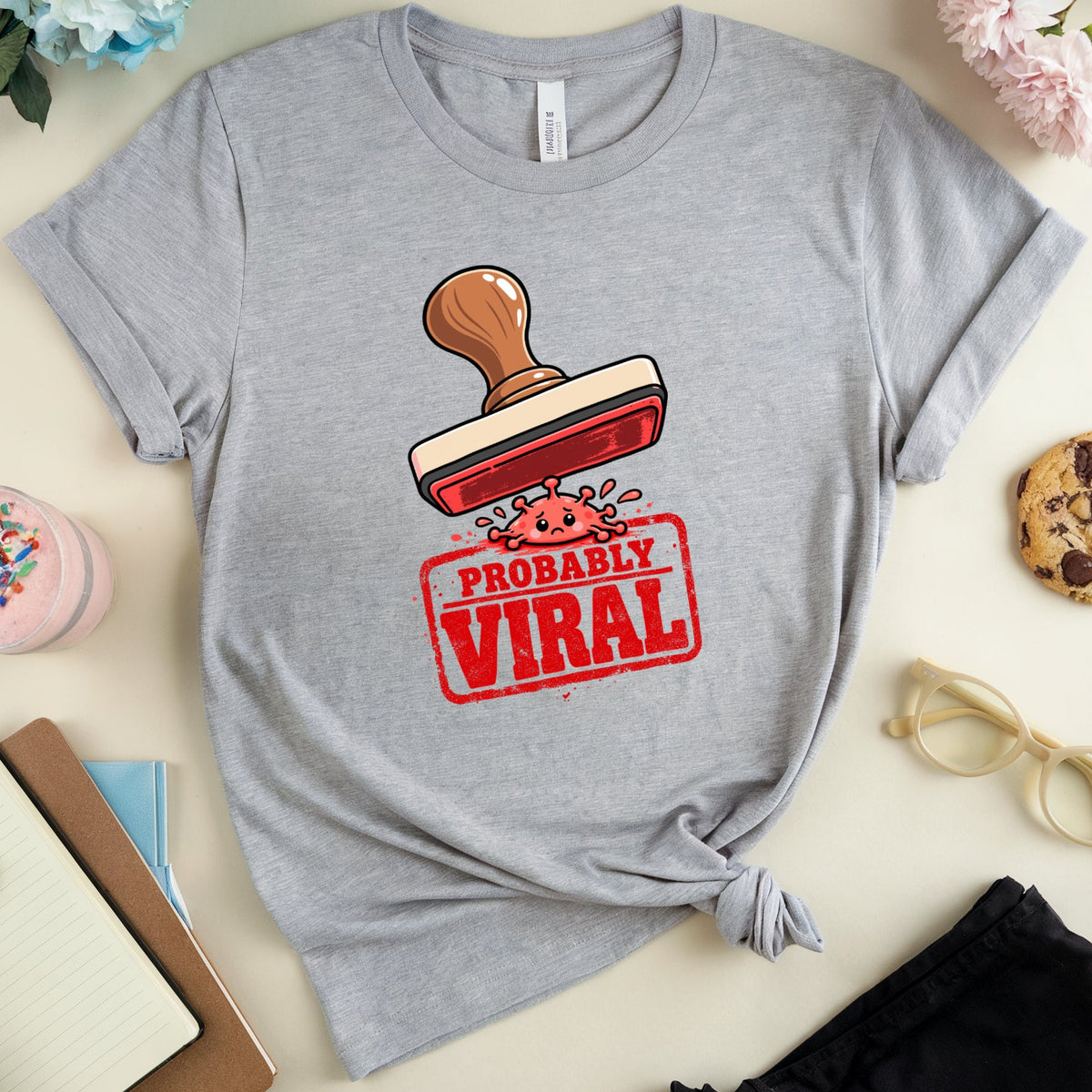 A heather gray short-sleeve T-shirt laid flat on a light beige surface, styled with flowers, a drink, a cookie, glasses, and notebooks around it. The shirt has a graphic of a red stamp with the words “PROBABLY VIRAL” and a small pink character.