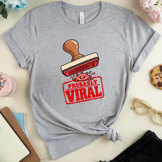 A heather gray short-sleeve T-shirt laid flat on a light beige surface, styled with flowers, a drink, a cookie, glasses, and notebooks around it. The shirt has a graphic of a red stamp with the words “PROBABLY VIRAL” and a small pink character.