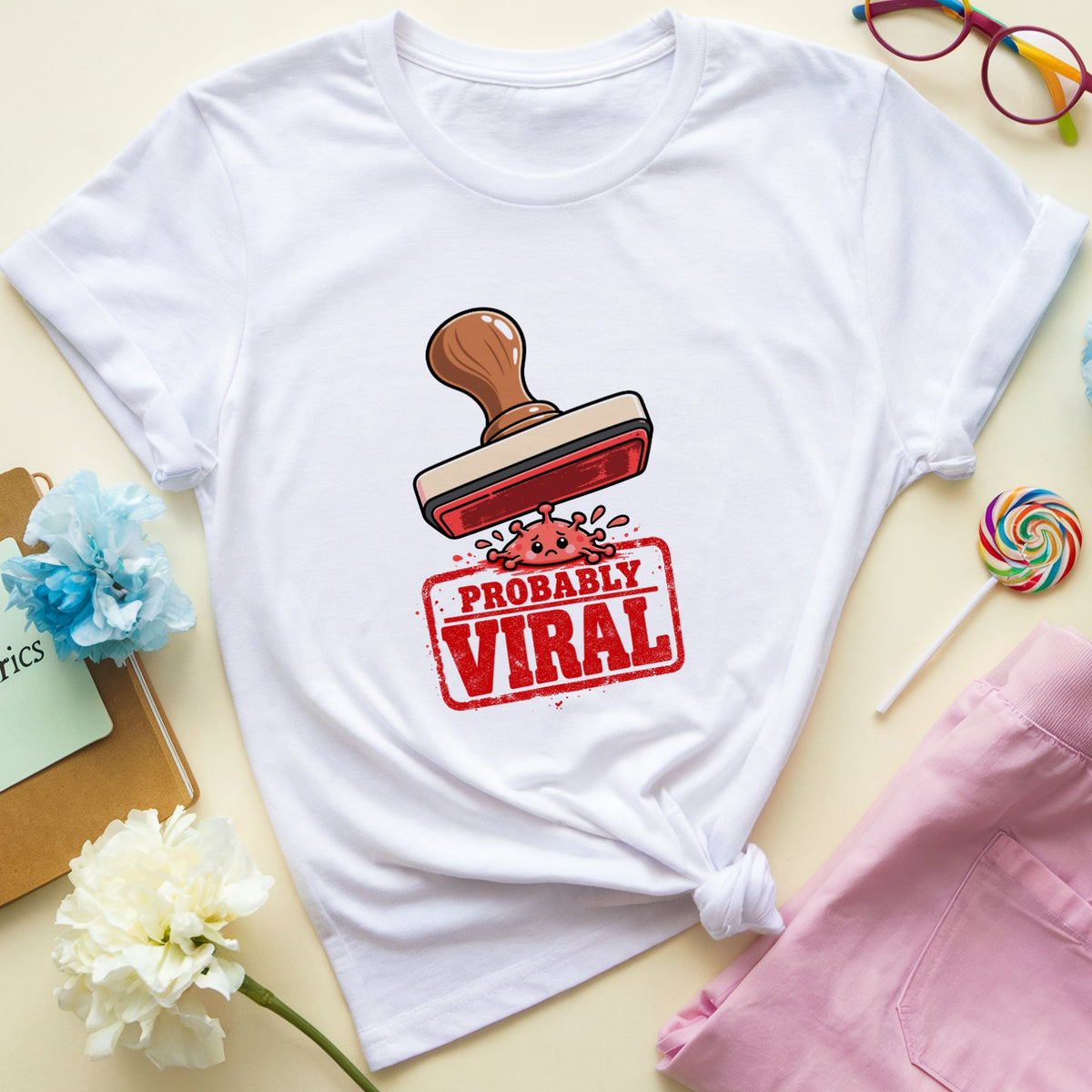 A white graphic t-shirt with a red and brown 'PROBABLY VIRAL' stamp design, styled in a flat lay with pink pajama pants, colorful glasses, a lollipop, flowers, and a notebook on a light beige background.