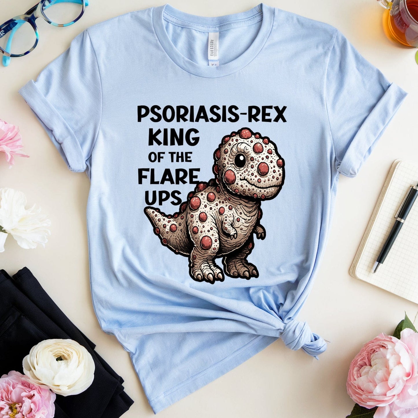 Light blue t-shirt with a dinosaur graphic and text on a table with flowers and a cup of tea.