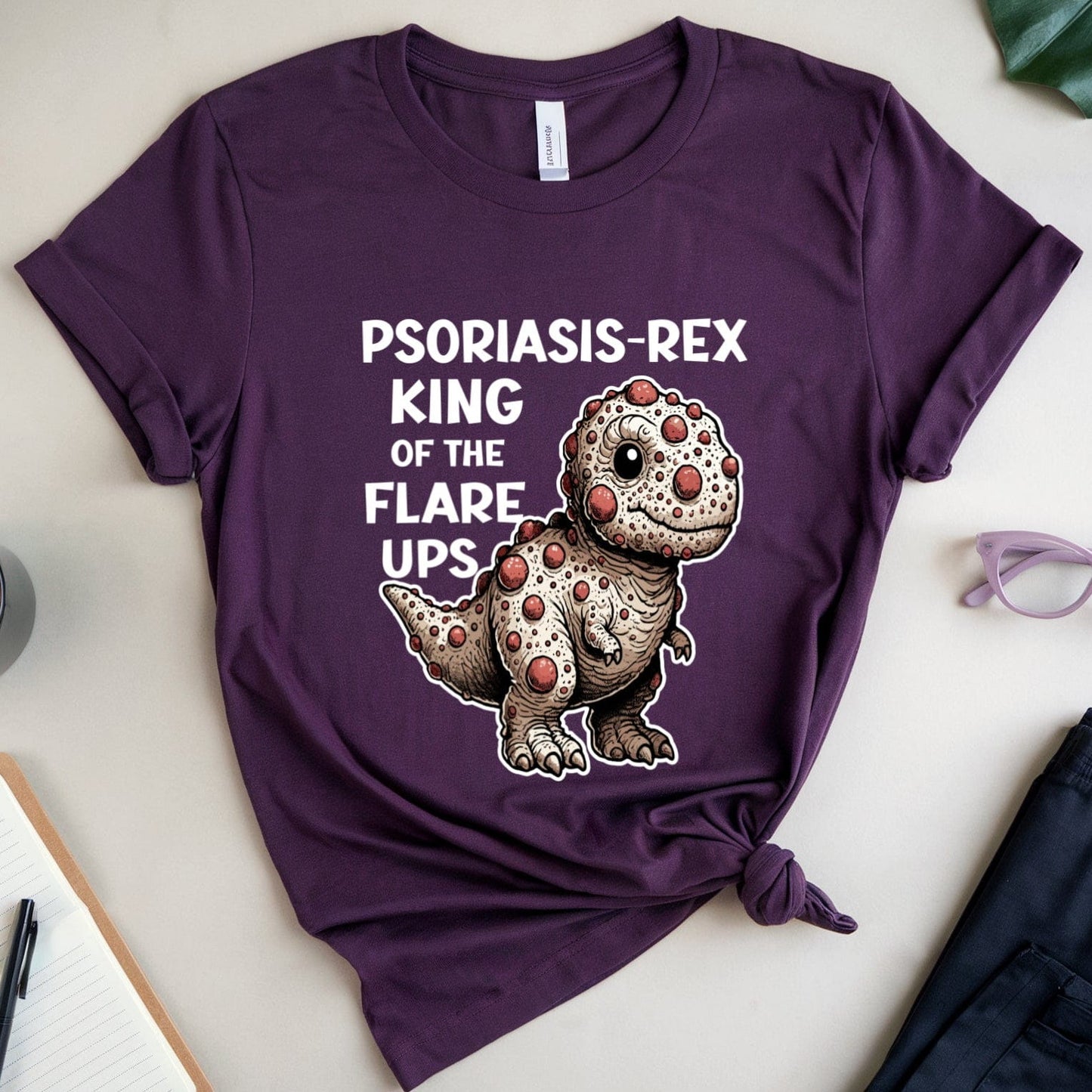 Purple t-shirt with dinosaur graphic and text on a light surface with a cup, notebook, and glasses.