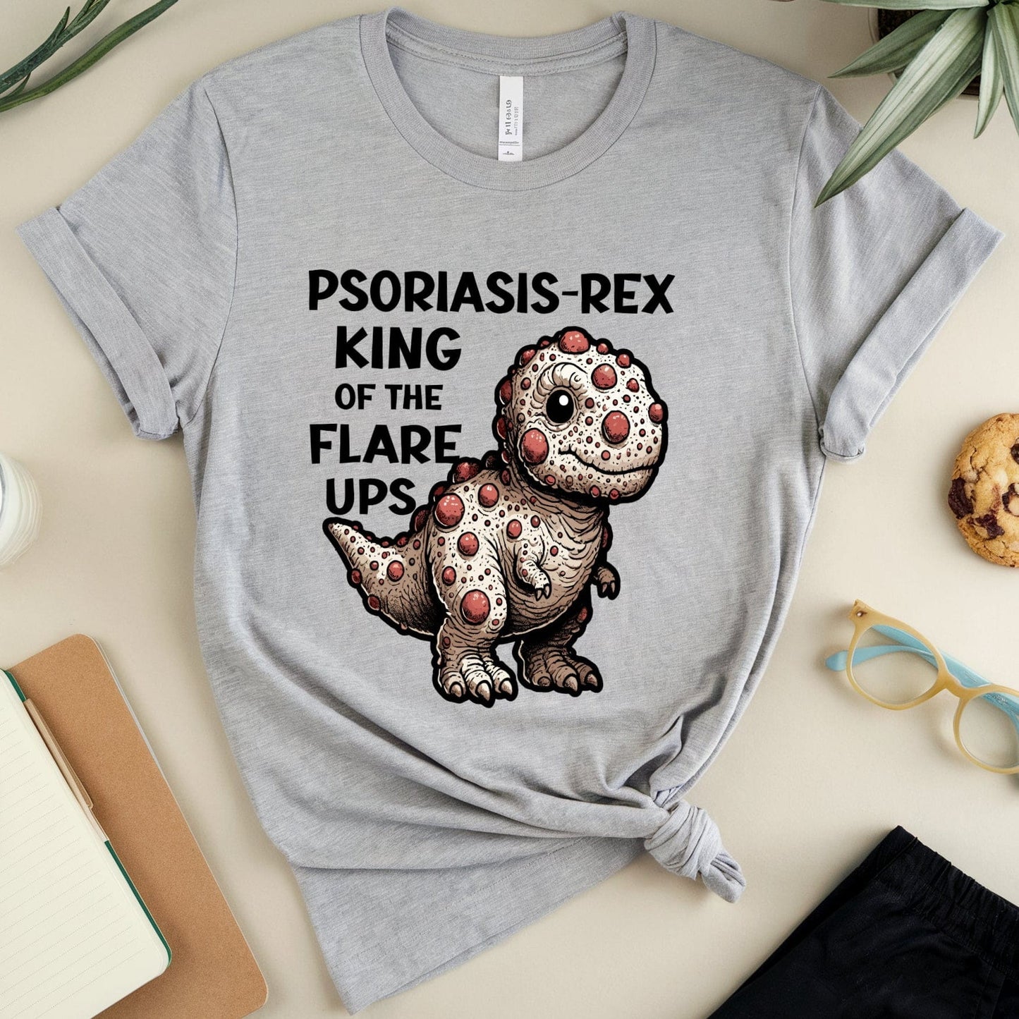 Gray t-shirt with a dinosaur graphic and text on a table with a glass, cookies, and glasses.