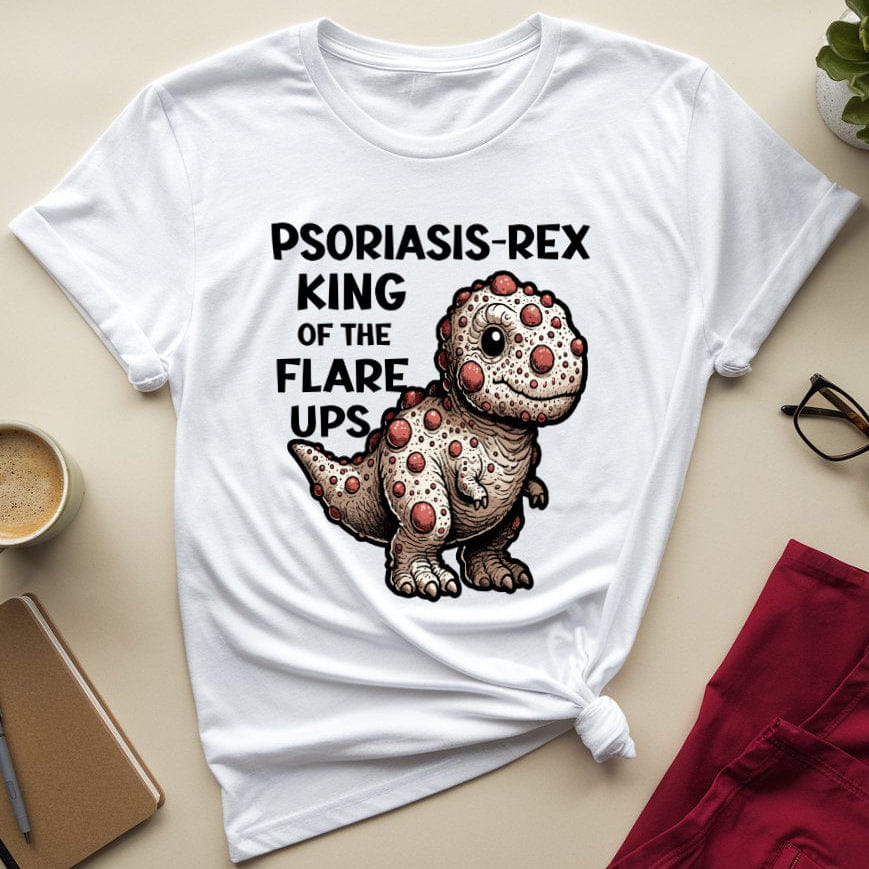 White t-shirt with a dinosaur graphic and text on a beige surface with a cup, glasses, and notebook.