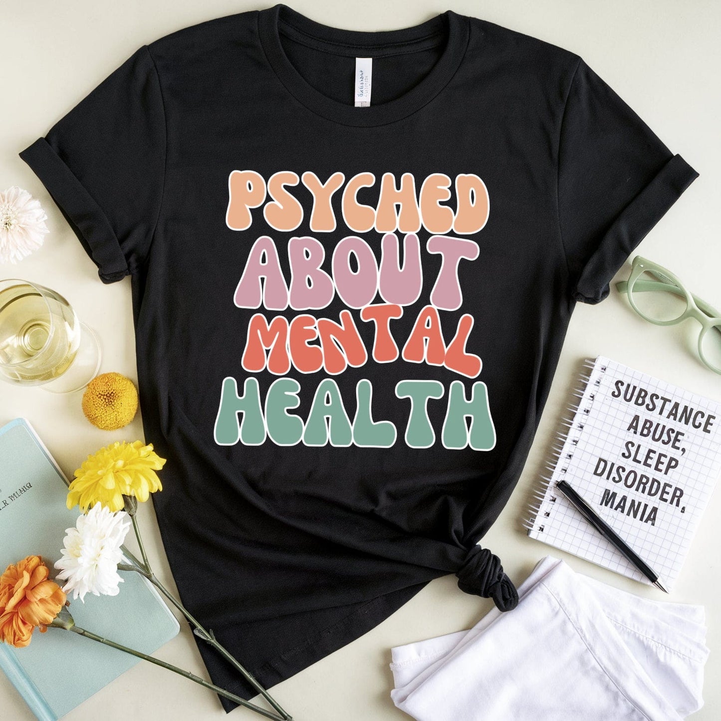 Black t-shirt with 'PSYCHED ABOUT MENTAL HEALTH' text on a light surface with flowers and books.