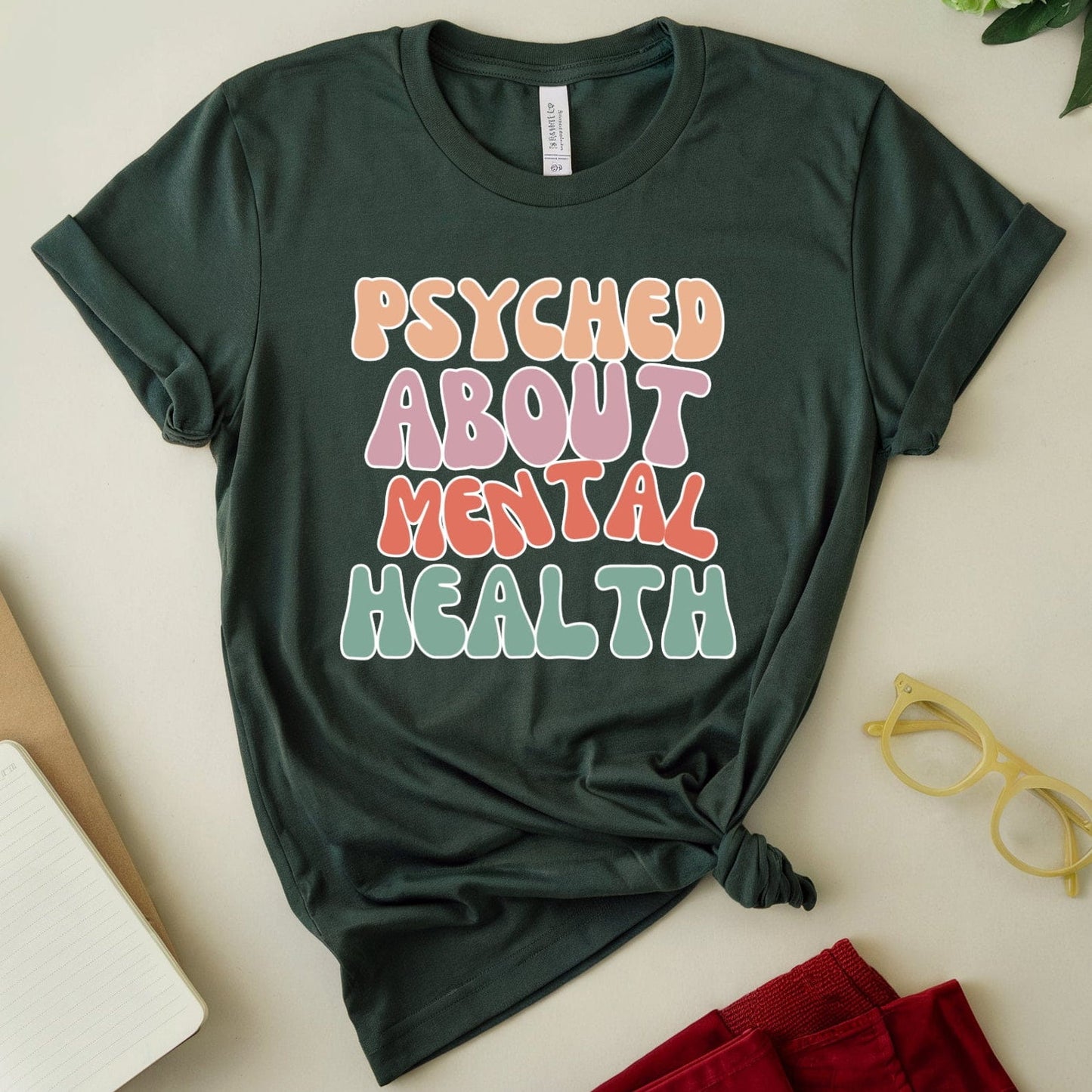 Green t-shirt with 'PSYCHED ABOUT MENTAL HEALTH' text on a light surface with accessories.