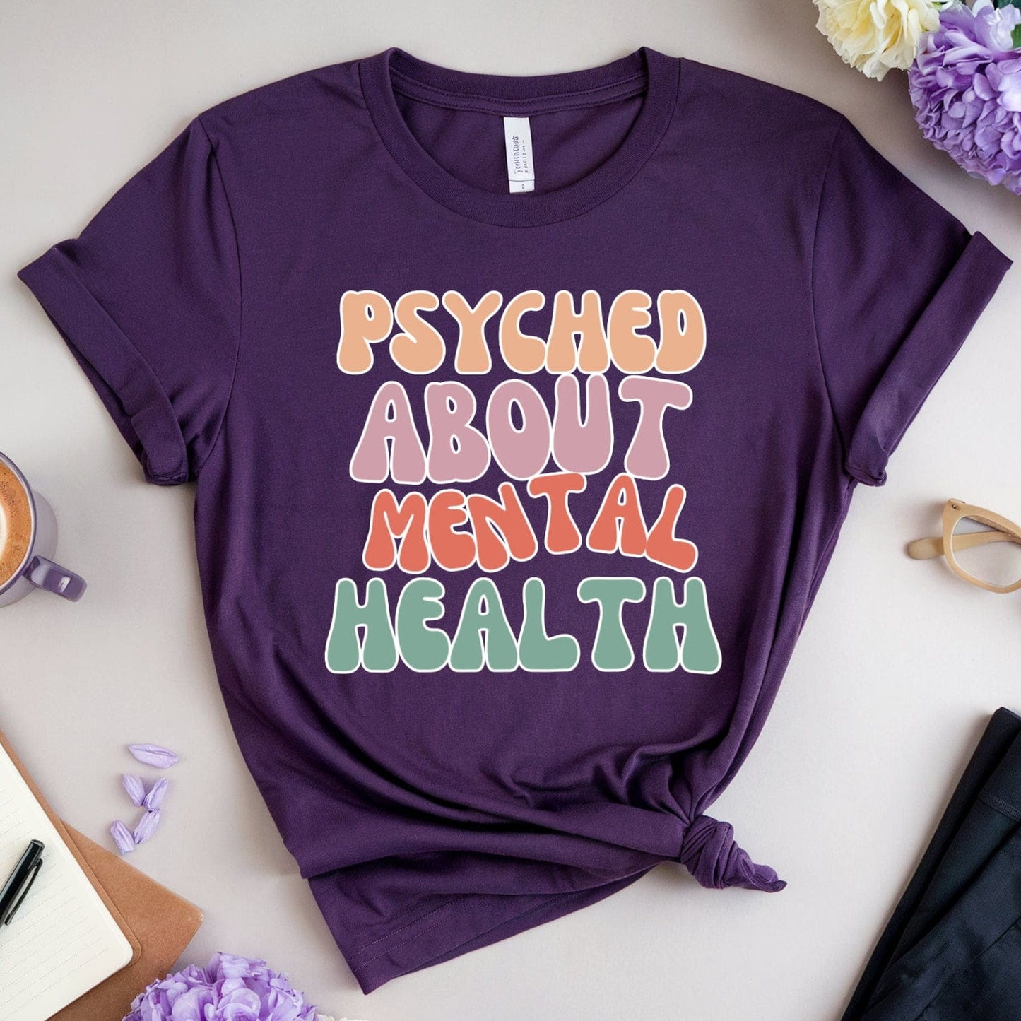 Purple t-shirt with 'PSYCHED ABOUT MENTAL HEALTH' text on a table with flowers and a cup of coffee.