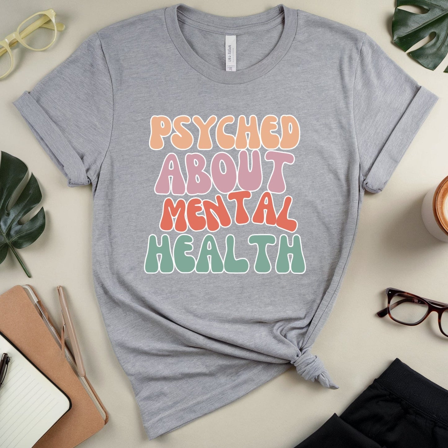Gray t-shirt with 'PSYCHED ABOUT MENTAL HEALTH' text on a light surface with accessories.