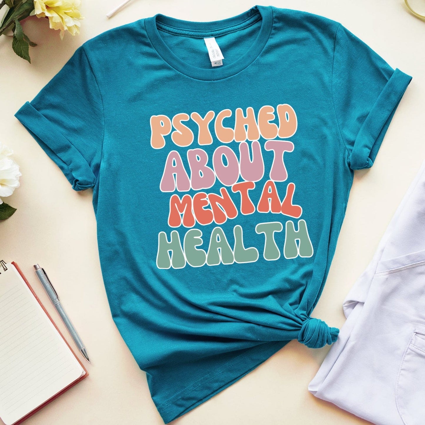 Blue t-shirt with 'Psyched About Mental Health' text on a light surface with flowers and glasses.