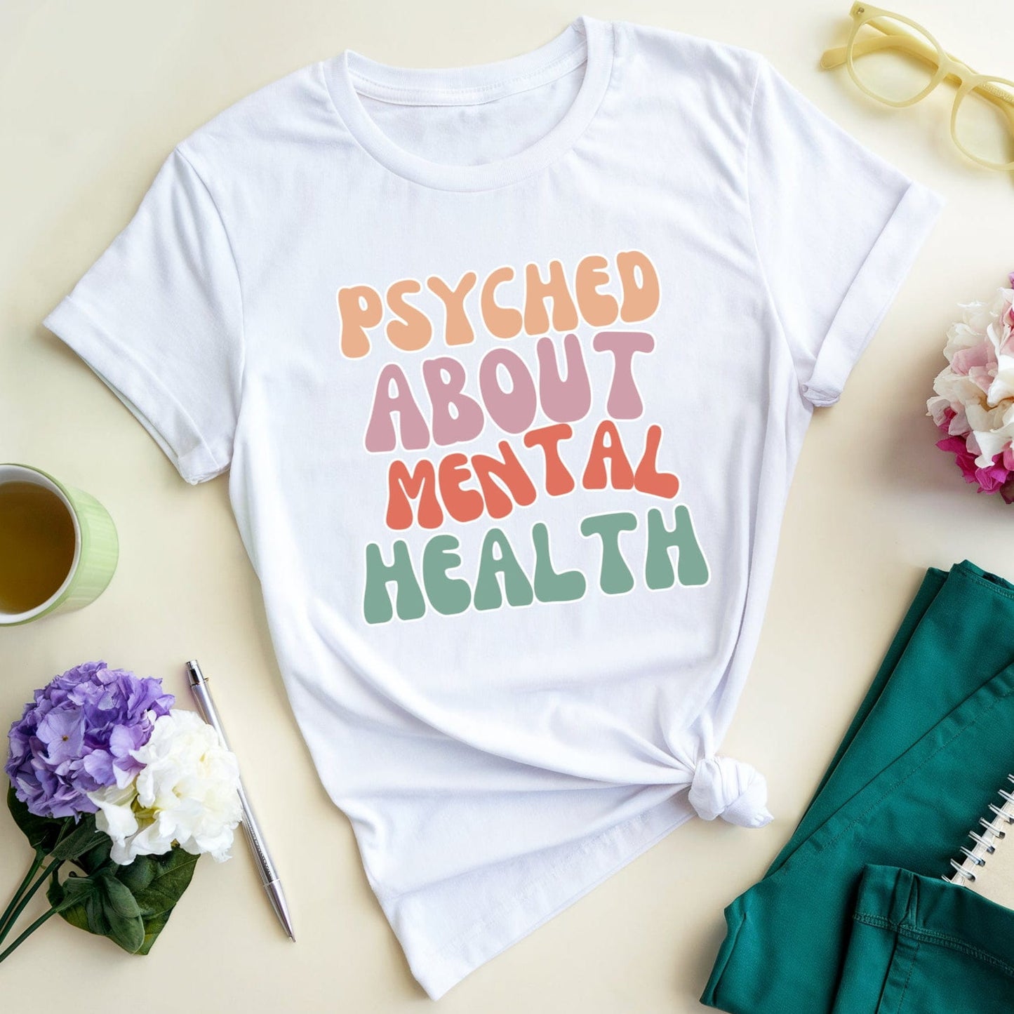White t-shirt with 'PSYCHED ABOUT MENTAL HEALTH' text on a light surface with flowers and a cup.
