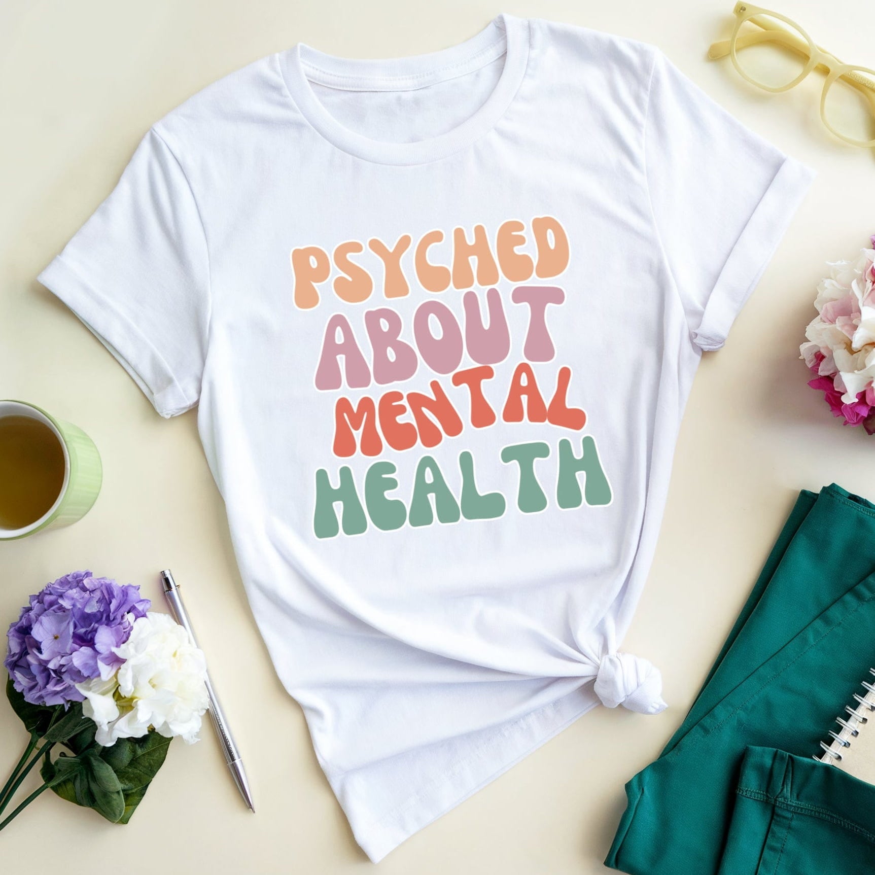 White t-shirt with 'PSYCHED ABOUT MENTAL HEALTH' text on a light surface with flowers and a cup.