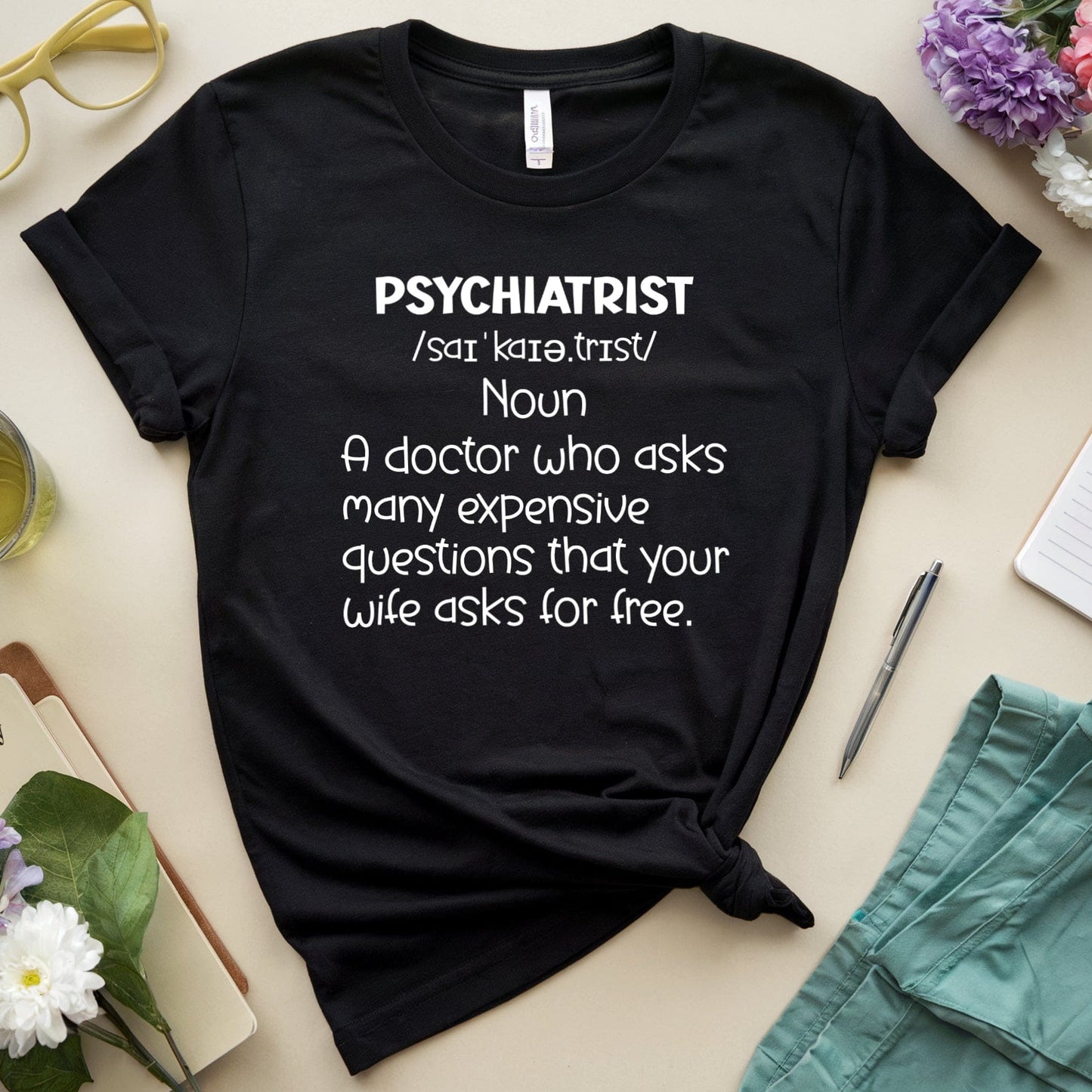 Black t-shirt with 'psychiatrist' definition text on a table with flowers and glasses.