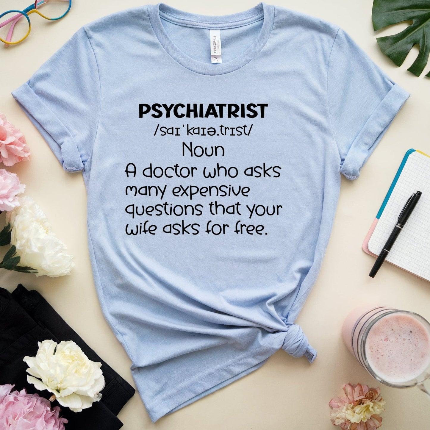 Light blue t-shirt with humorous definition of a psychiatrist text, placed on a surface with flowers, glasses, and a notebook.