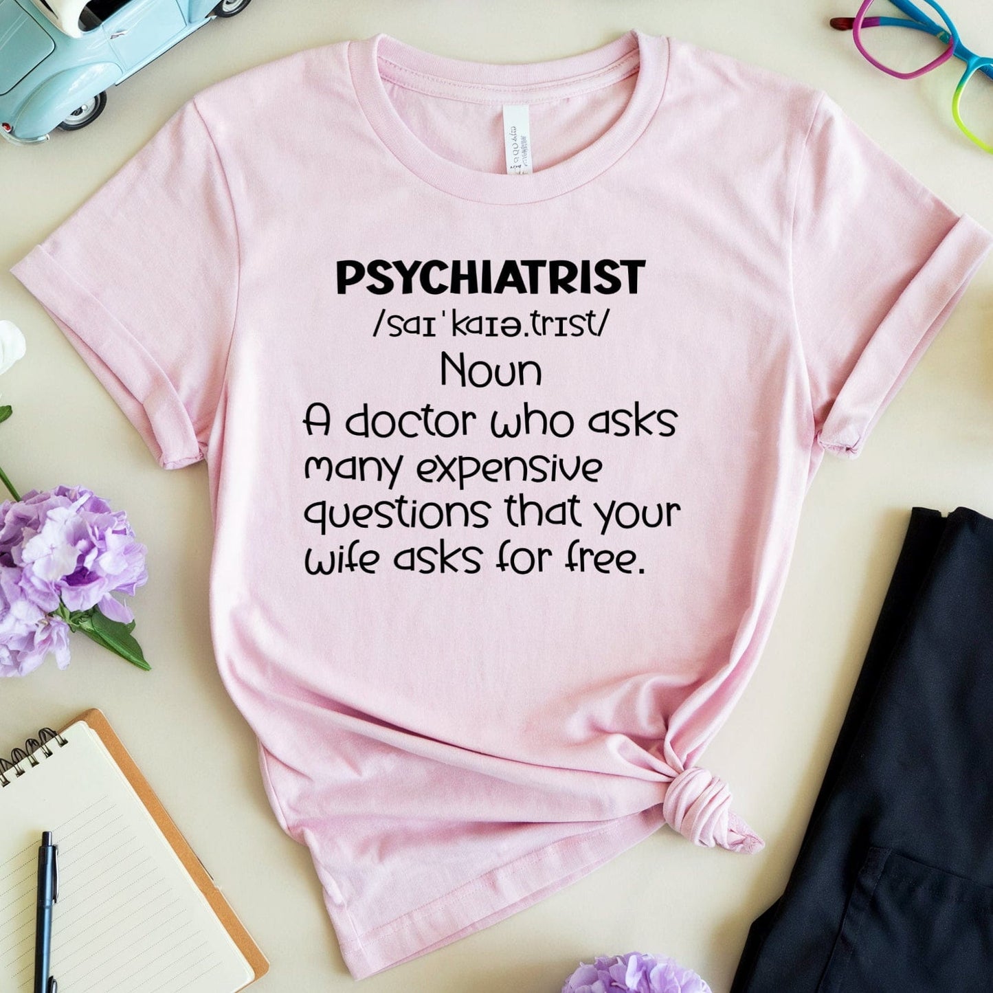 Pink t-shirt with 'psychiatrist' definition text on a light surface with flowers and a toy car.