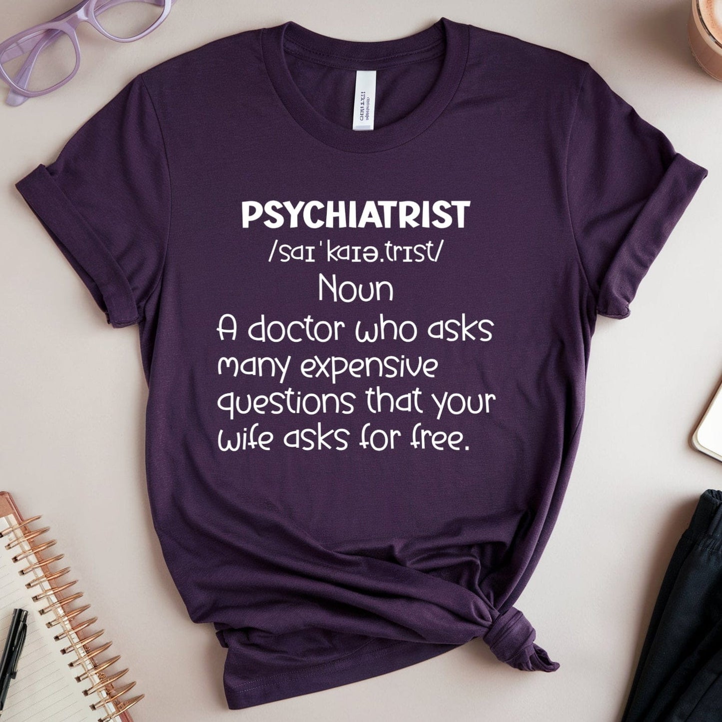 Purple t-shirt with humorous definition of a psychiatrist on a table with glasses, notebook, and coffee.