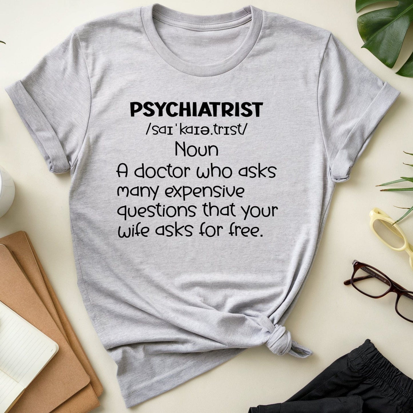 Gray t-shirt with humorous psychiatrist definition text on a desk with office items.
