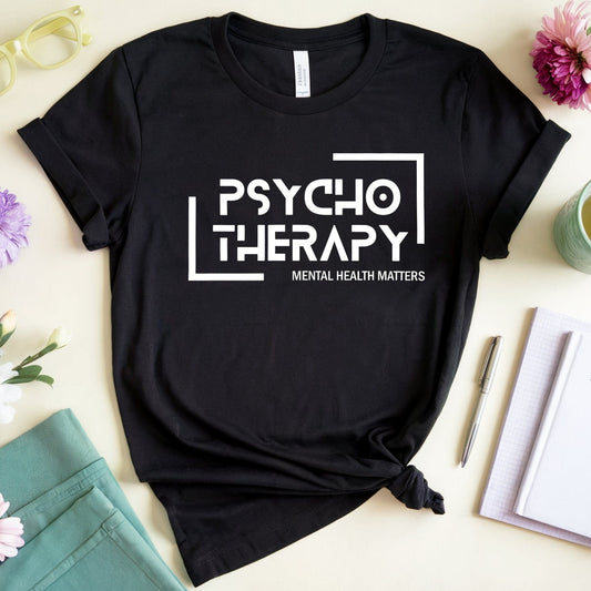 Black t-shirt with 'Psycho Therapy' text on a table with flowers and a notebook.