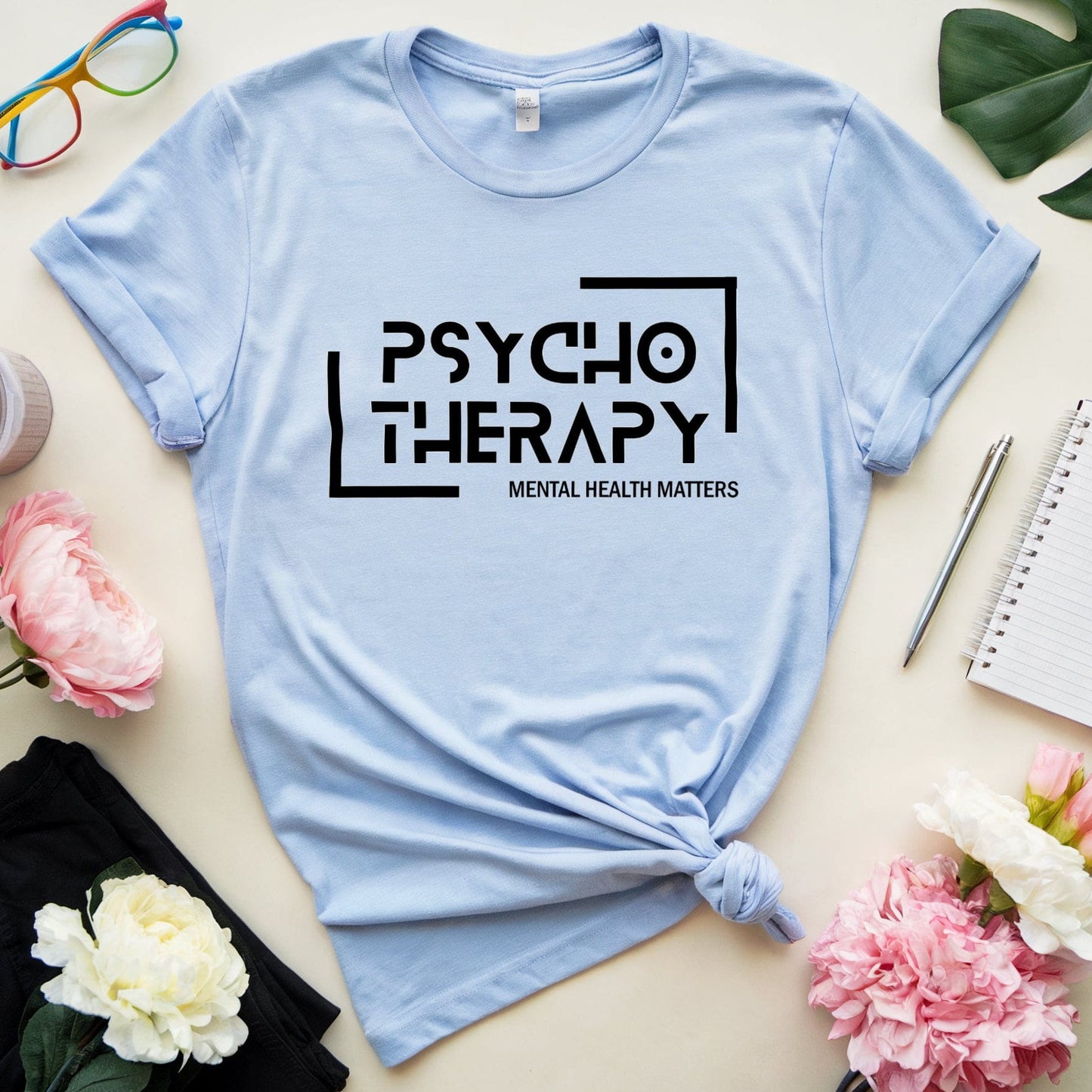Light blue t-shirt with 'Psycho Therapy' text on a table with flowers and glasses.