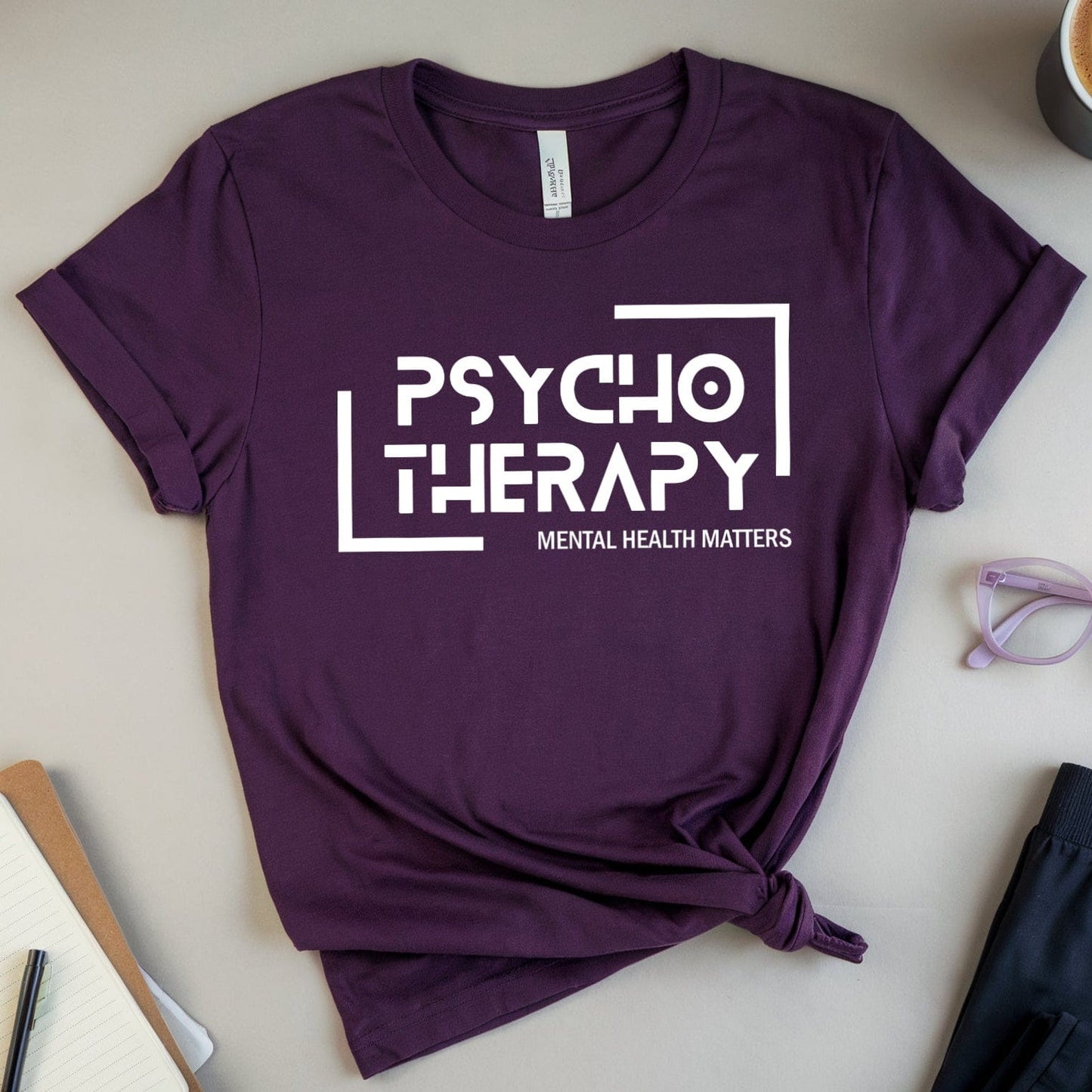 Purple t-shirt with 'Psychotherapy Mental Health Matters' text on a light surface with a cup of coffee, glasses, and books.