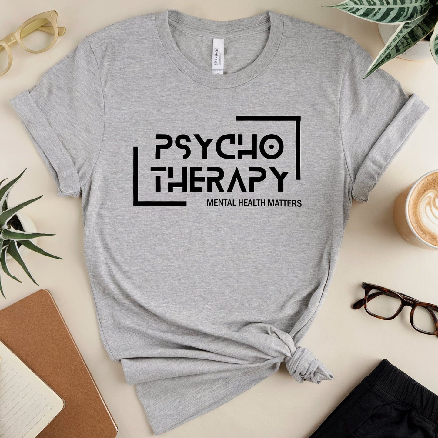Gray t-shirt with 'Psychotherapy Mental Health Matters' text on a table with glasses, notebook, and plant.