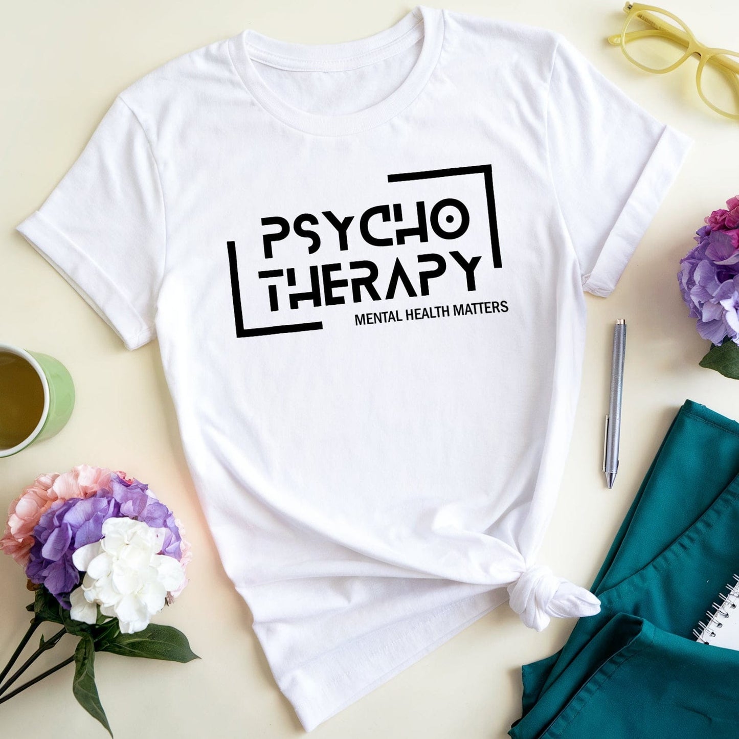 White t-shirt with 'Psycho Therapy' text on a table with flowers, cup, and notebook.