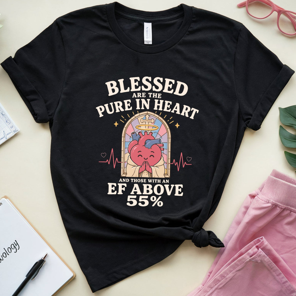 A black graphic t-shirt is displayed in a styled flat-lay arrangement on a light beige background. The shirt features a graphic of a smiling red heart inside a stained-glass window with the text 'BLESSED ARE THE PURE IN HEART AND THOSE WHO SEE EF ABOVE 55%'.