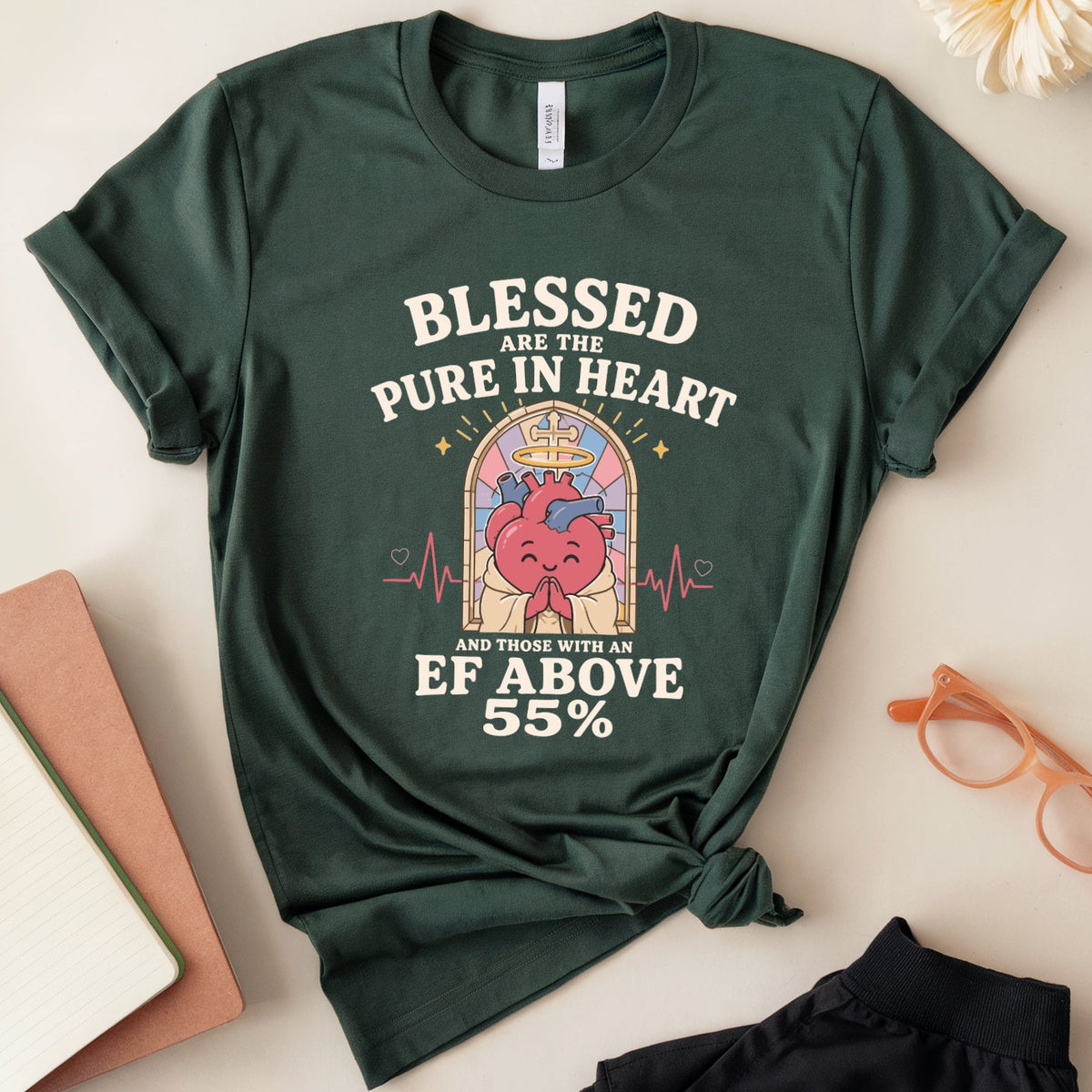 A dark green graphic t-shirt with a heart-shaped stained glass design and the text 'BLESSED ARE THE PURE IN HEART AND THOSE WITH AN EF ABOVE 55%'. The t-shirt is styled in a flat-lay arrangement on a light beige background with a notebook, pen, orange glasses, black pants, and a white flower.