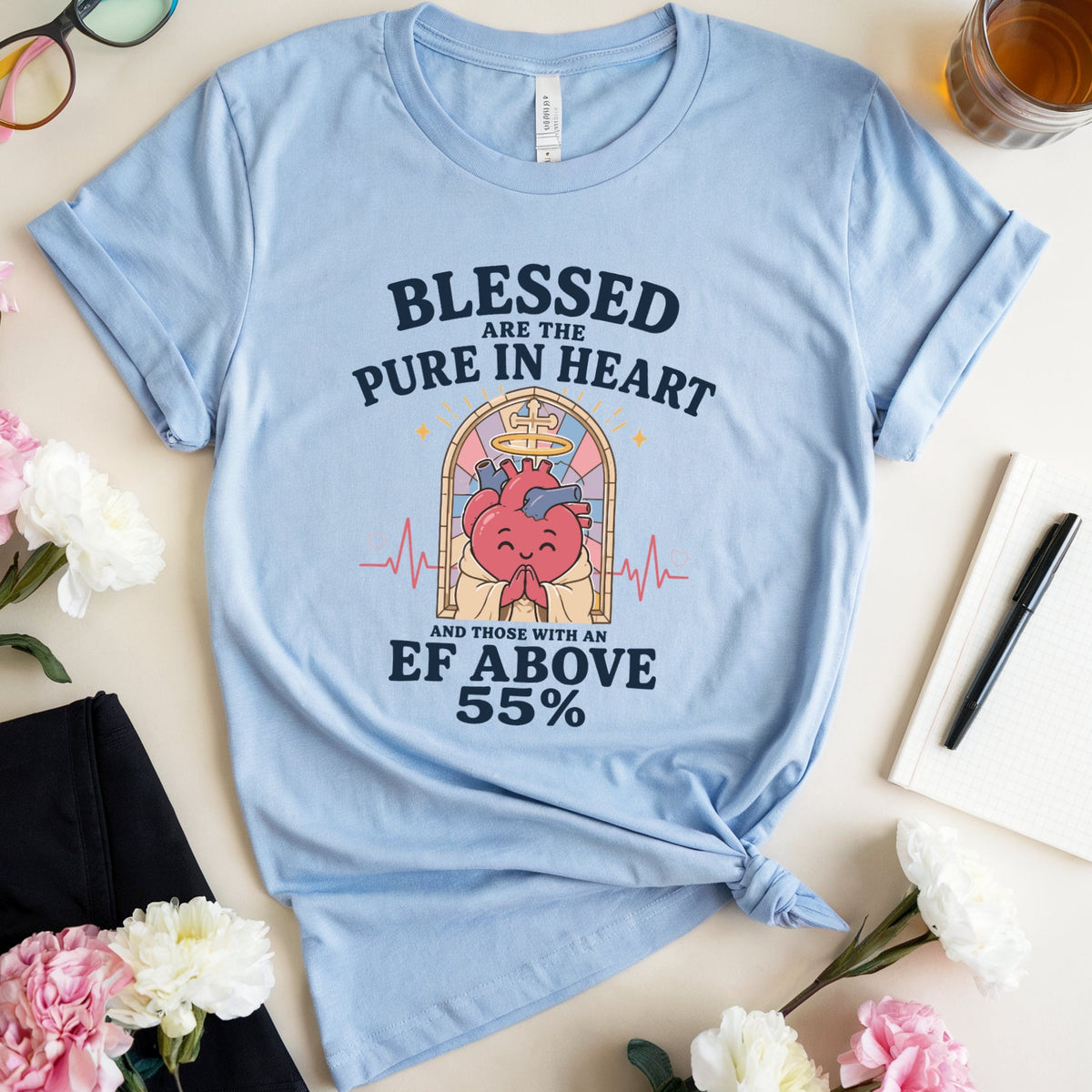 A light blue graphic t-shirt is styled in a flat lay on a beige surface. The t-shirt features a graphic of a cartoon heart with a crown and a pink-and-blue heart, surrounded by text that reads 'BLESSED ARE THE PURE IN HEART AND THOSE WITH AN EF ABOVE 55%'.