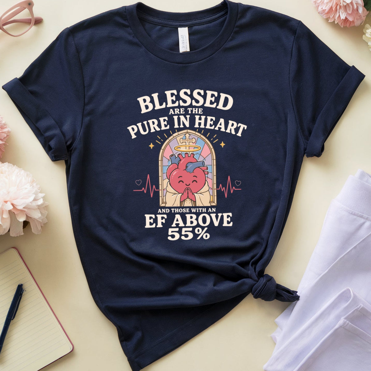 A navy blue graphic t-shirt with a heart design and the text 'BLESSED ARE THE PURE IN HEART EF ABOVE 55' is styled in a flat lay with white pants, pink flowers, glasses, a notebook, and a cup on a light beige background.