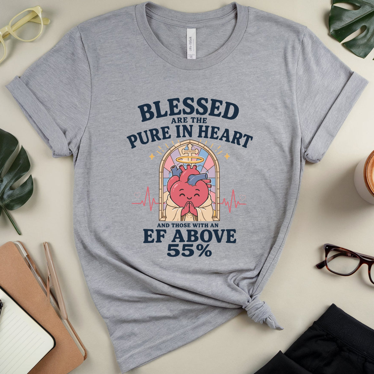 A heather gray graphic t-shirt with a heart-shaped illustration and the text 'Blessed are the Pure in Heart and those with an EF above 55%' displayed in a styled flat-lay with glasses, leaves, a notebook, and a coffee cup.