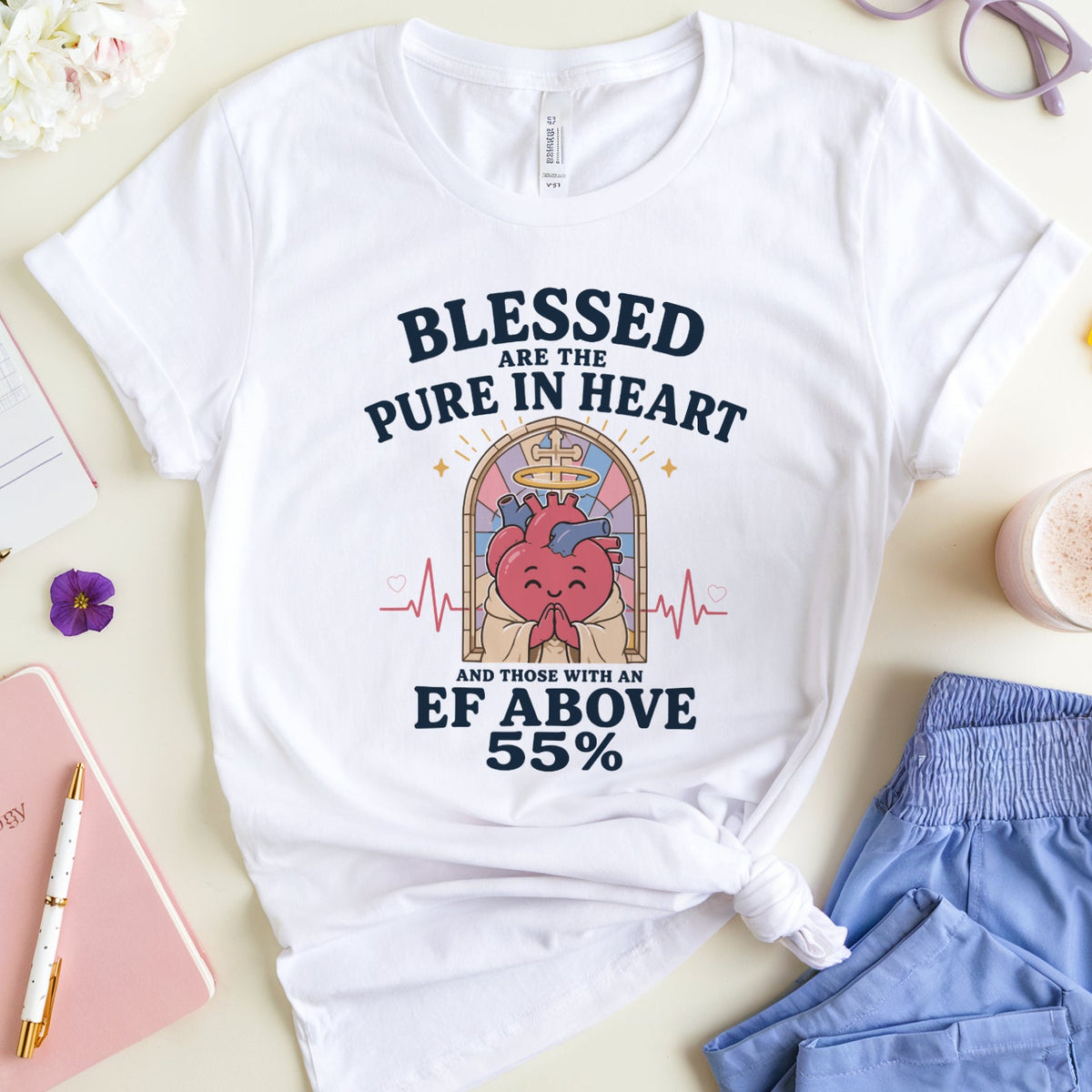 A white graphic T-shirt laid flat on a light beige surface, surrounded by styled props including flowers, glasses, a notebook, a pink drink, and blue shorts. The shirt has a large printed slogan and heart illustration on the front.