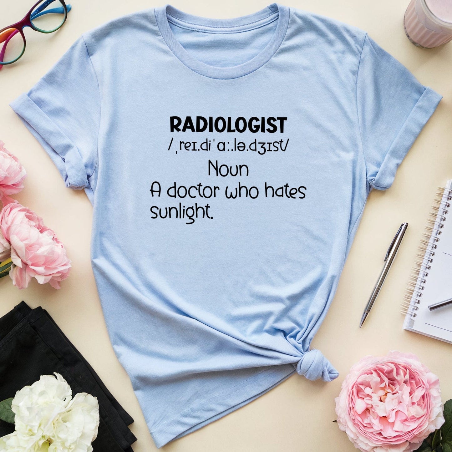 Light blue t-shirt with humorous radiologist definition on a table with flowers and glasses.