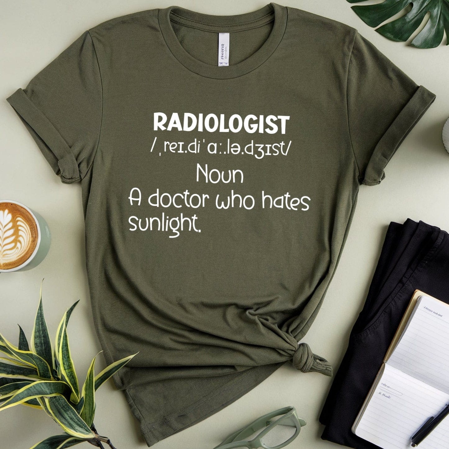 Green t-shirt with 'Radiologist' definition text on a light surface with plants and a cup.