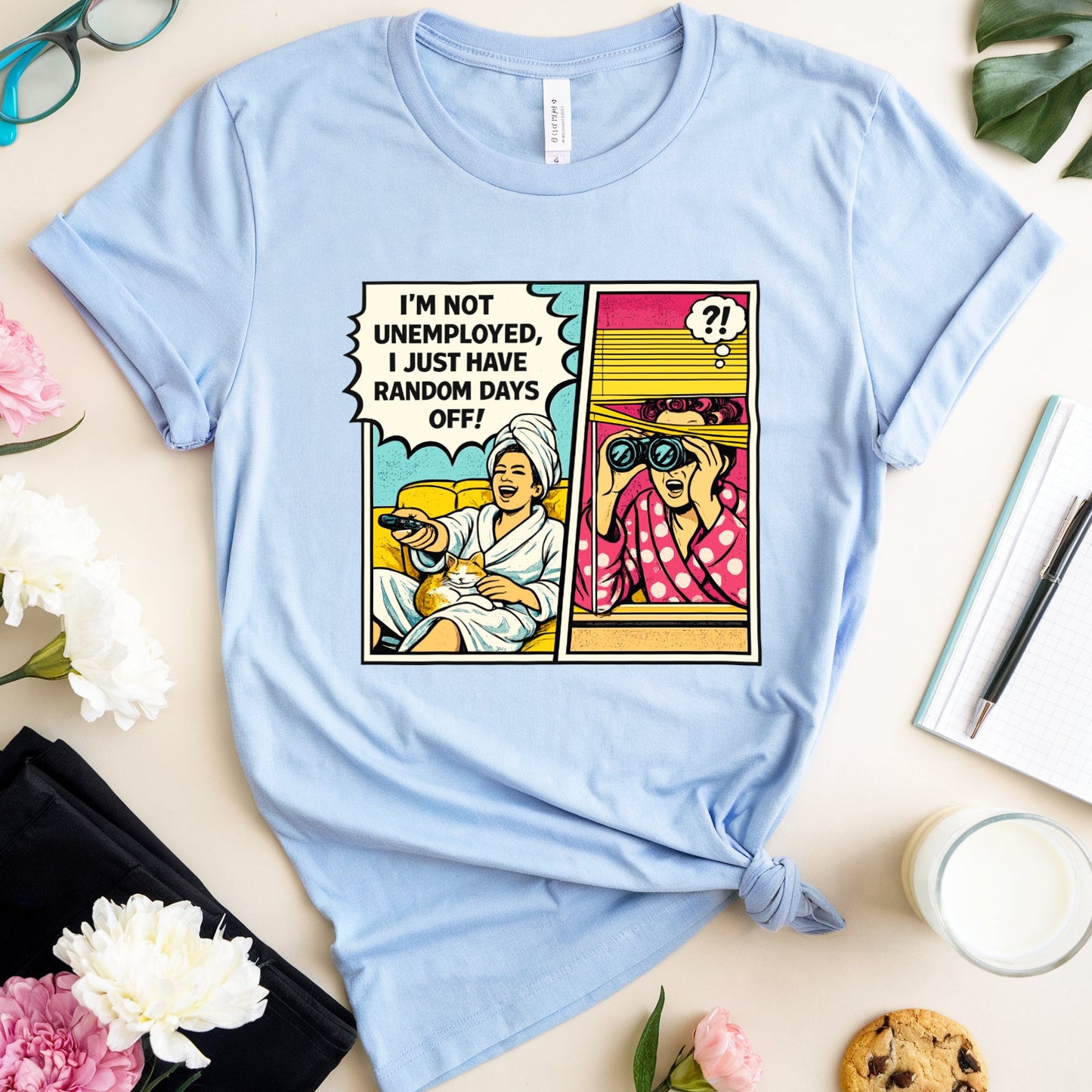 Light blue t-shirt with a graphic design on a table with flowers and a glass of milk.