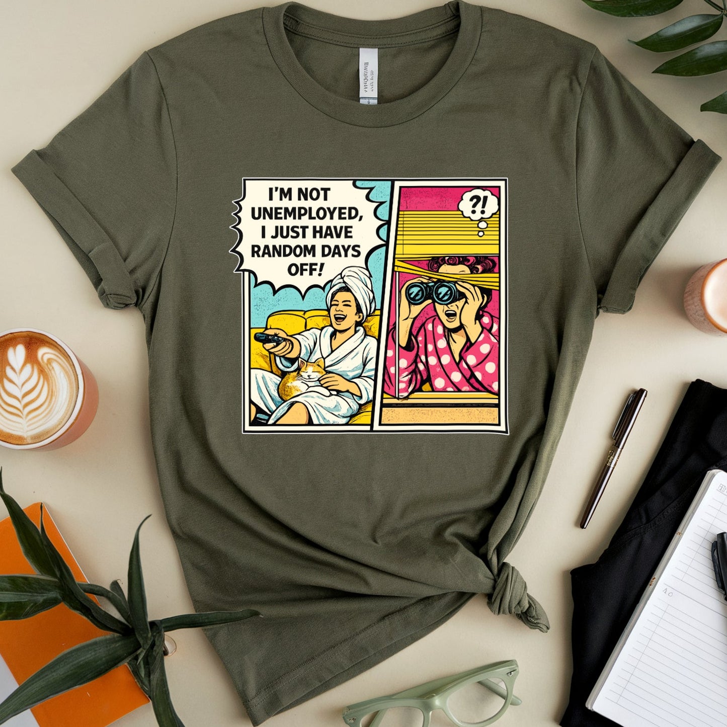 Green t-shirt with a graphic design on a table with a cup, glasses, and notebook.