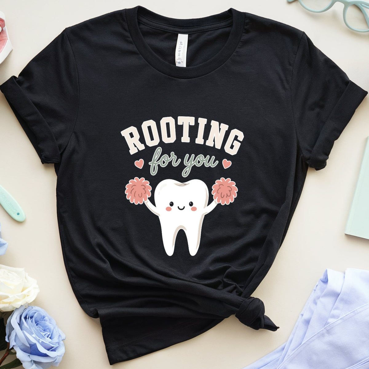 A black graphic T-shirt laid flat on a beige surface, surrounded by dental-themed props including a tooth model, mouth model, toothbrush, glasses, flowers, a notebook, and folded scrubs. The shirt has a cartoon tooth holding pink pom-poms with the text 'ROOTING for you'.