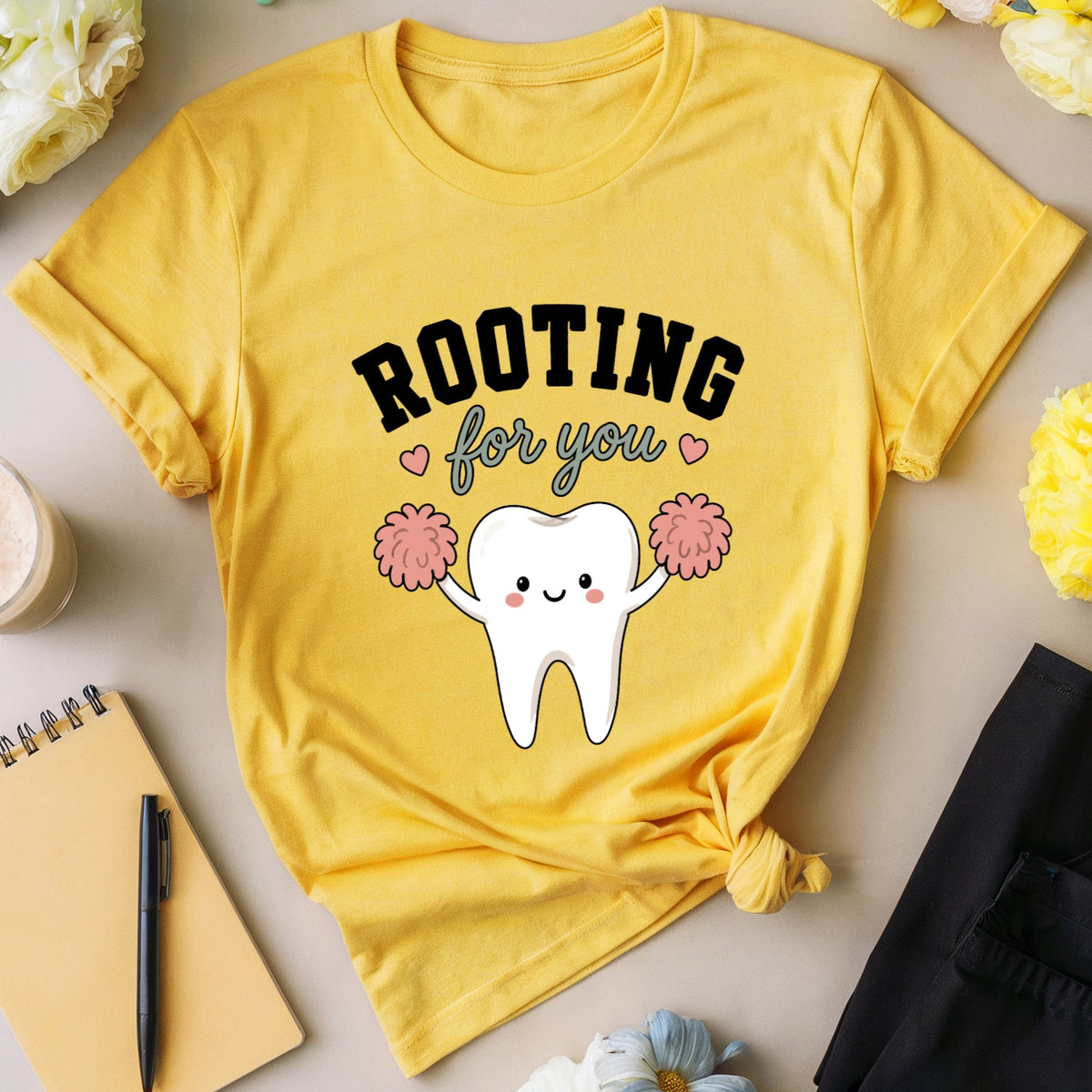 A yellow graphic T-shirt laid flat on a beige surface, surrounded by flowers, eyeglasses, a drink, a notebook, pen, and folded black pants. The shirt has a cartoon tooth with pink pom-poms and the text 'ROOTING for you' printed on the front.