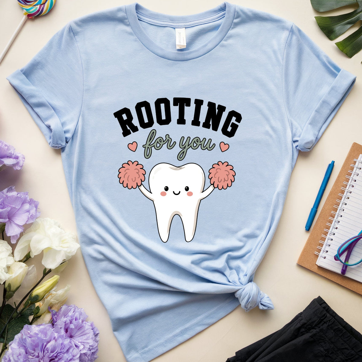 A light blue graphic t-shirt with a 'Rooting for you' cartoon tooth cheerleader design, styled in a flat lay with flowers, a lollipop, glasses, a notebook, and a plant leaf on a beige background.