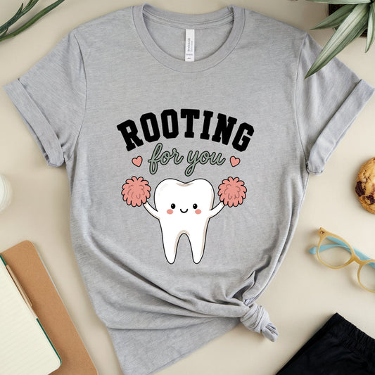 A heather gray short-sleeve T-shirt laid flat on a light beige surface, styled with a tooth graphic that reads “ROOTING for you” with a smiling cartoon tooth holding pink pom-poms. The shirt is tied at the hem and surrounded by props including a notebook, pen, cookie, eyeglasses, shorts, a glass of milk, and plants.