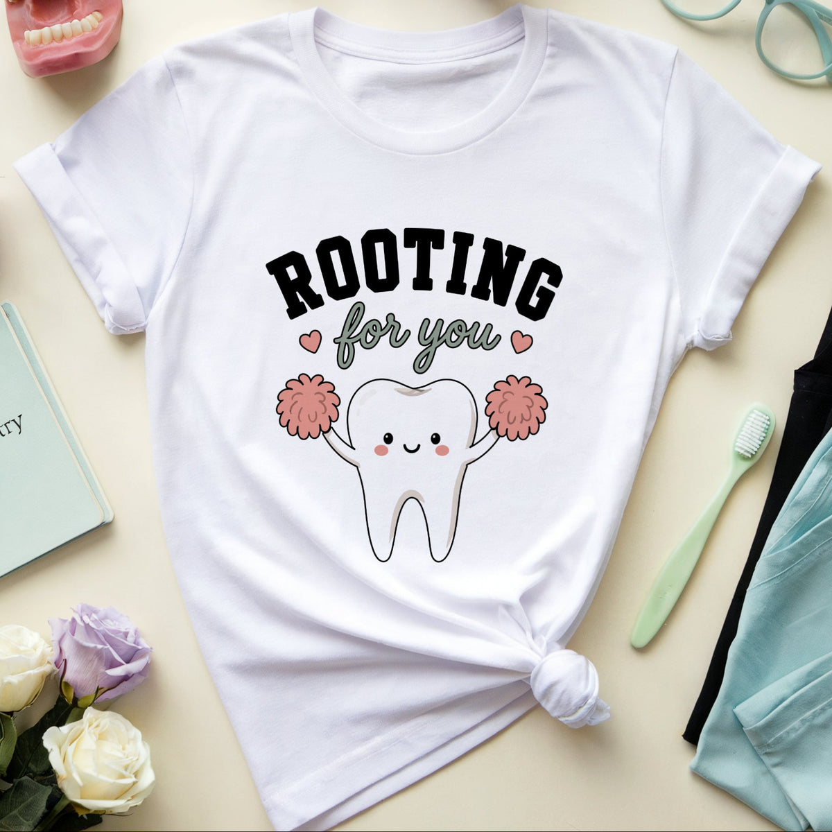 A white graphic T-shirt laid flat on a light beige surface, surrounded by dental-themed props including a model teeth mouth, purple and white roses, eyeglasses, a notebook, a toothbrush, and folded scrubs. The shirt graphic reads “ROOTING for you” with a cute cartoon tooth holding pink pom-poms.