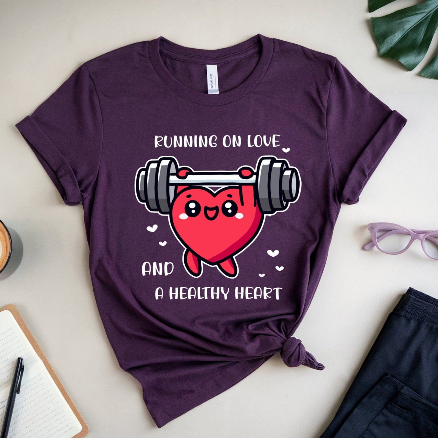 Purple t-shirt with heart and dumbbell graphic and text 'Running on love and a healthy heart' on a light surface with coffee, glasses, and shorts.