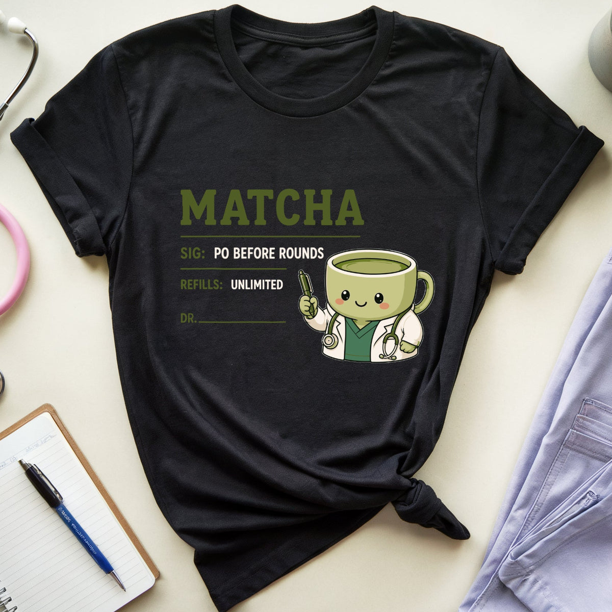 A black graphic T-shirt laid flat on a light beige surface, styled with a pink stethoscope, notebook, pen, folded lavender scrubs, and a coffee mug. The shirt has a matcha-themed medical badge graphic with the word MATCHA in olive green and a cute matcha cup character holding a syringe.