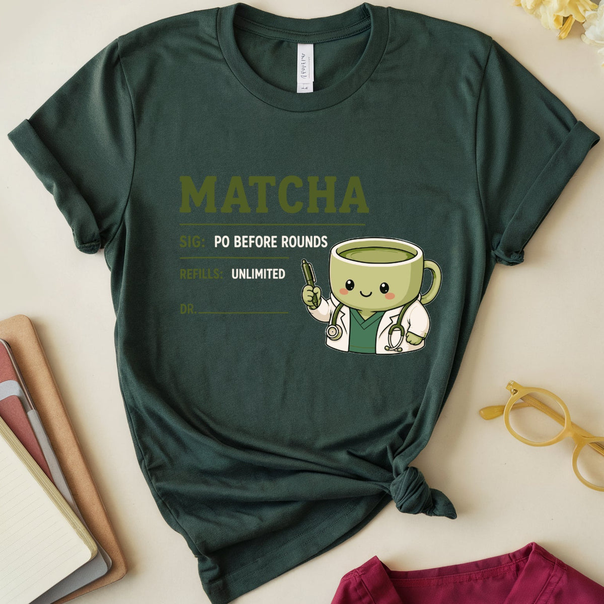 A dark green graphic t-shirt featuring a cute matcha doctor cartoon and the text 'MATCHA PO BEFORE ROUNDS', styled in a flat lay with notebooks, glasses, and flowers on a light beige background.
