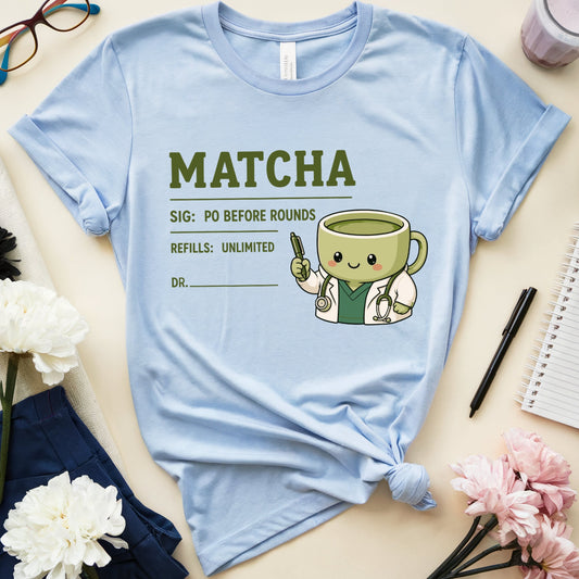 A light blue graphic T-shirt laid flat on a beige surface, styled with flowers, glasses, a notebook, and a drink. The shirt has a matcha-themed print reading "MATCHA" with a cute smiling matcha cup character and text below.