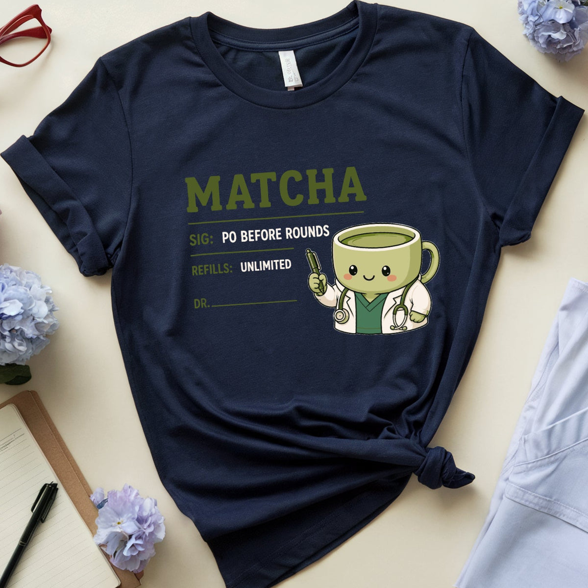 A navy blue graphic T-shirt laid flat on a light beige surface, styled with flowers, red eyeglasses, an open notebook, a coffee mug, and folded light blue scrubs. The shirt has a green 'MATCHA' graphic and a cartoon matcha cup with a stethoscope.