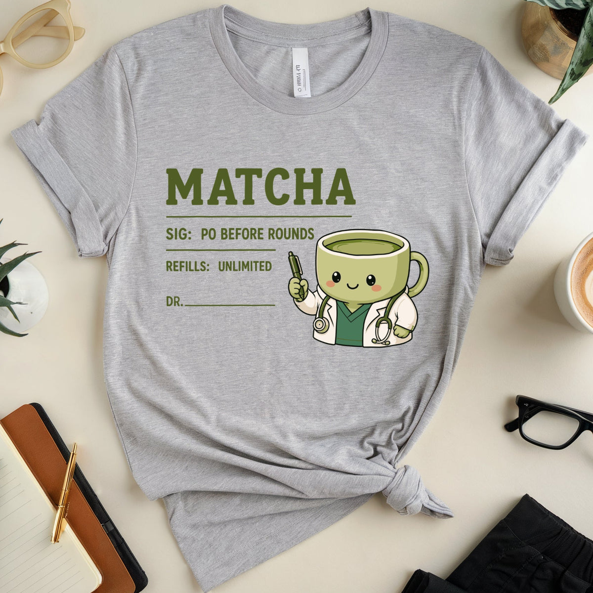 A heather gray graphic T-shirt laid flat on a light beige surface, surrounded by lifestyle props including a cup of latte art coffee, eyeglasses, notebooks, and plants. The shirt features a matcha-themed medical-style print with the word 'MATCHA' and a cute green tea cup character holding a matcha leaf.